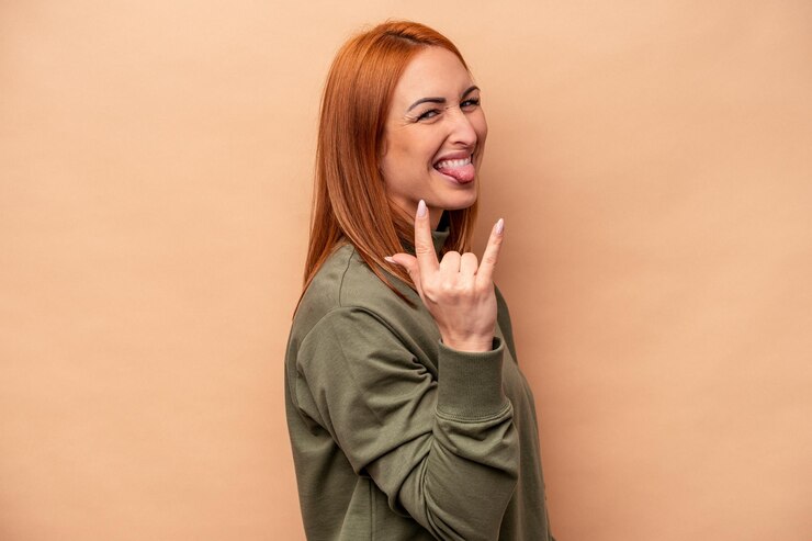 Giovane donna caucasica isolata su fondo beige che mostra gesto rock con le dita