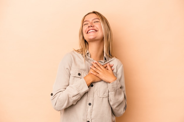 Giovane donna caucasica isolata su fondo beige che ride tenendo le mani sul cuore, concetto di felicità.