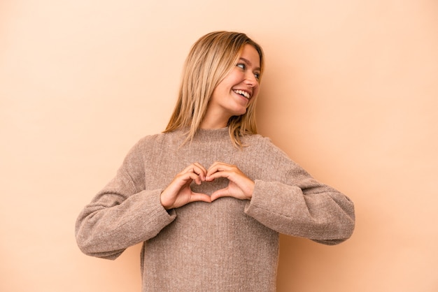 Giovane donna caucasica isolata su fondo beige che sorride e che mostra una forma del cuore con le mani.