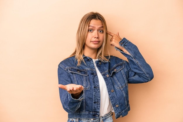 Giovane donna caucasica isolata su fondo beige che tiene e mostra un prodotto a portata di mano.