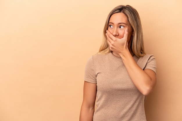 Giovane donna caucasica isolata su fondo beige premurosa che guarda ad uno spazio della copia che copre la bocca con la mano.