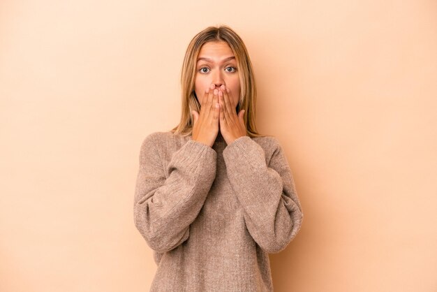 Giovane donna caucasica isolata su fondo beige scioccata che copre la bocca con le mani.