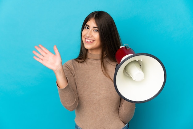 Giovane donna caucasica isolata su priorità bassa blu che tiene un megafono e che saluta con la mano con l'espressione felice