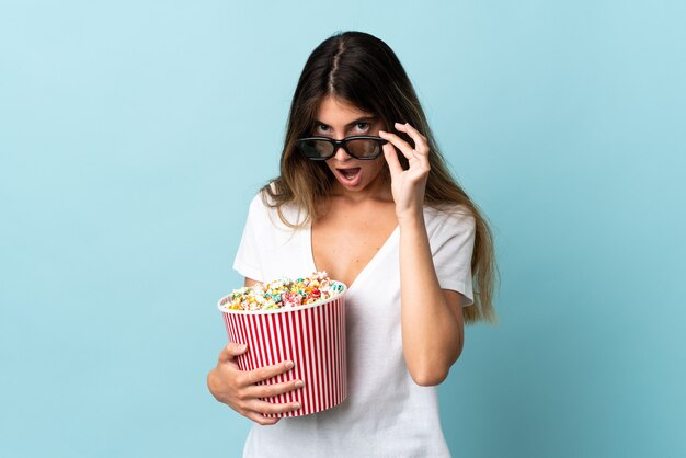 Giovane donna caucasica isolata su priorità bassa blu sorpresa con gli occhiali 3d e che tiene un grande secchio di popcorn