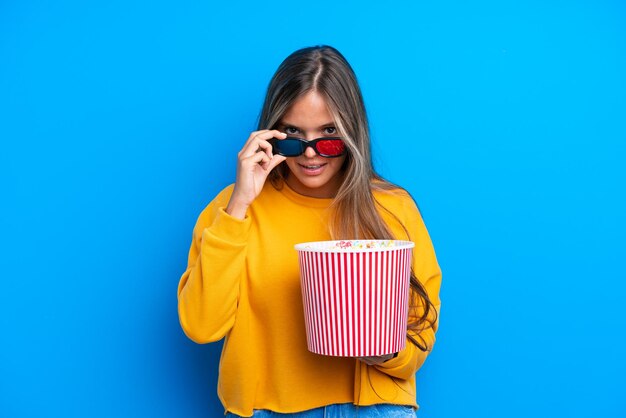 Giovane donna caucasica isolata su priorità bassa blu sorpresa con gli occhiali 3d e che tiene un grande secchio di popcorn