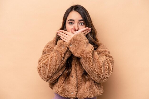 Giovane donna caucasica isolata su sfondo beige che copre la bocca con le mani che sembrano preoccupate