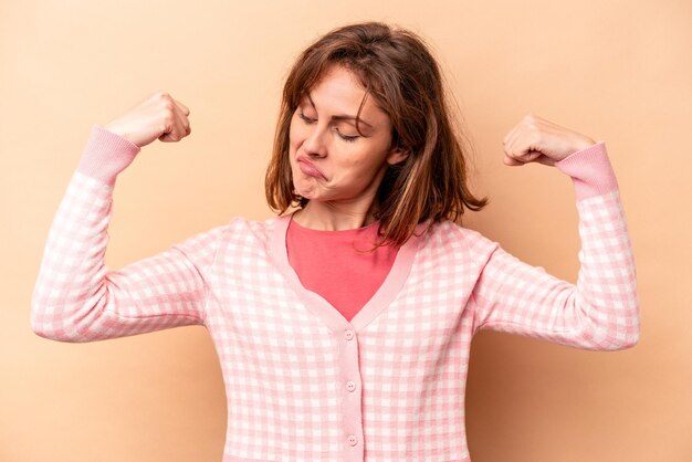 Giovane donna caucasica isolata su sfondo beige che mostra gesto di forza con le braccia simbolo del potere femminile