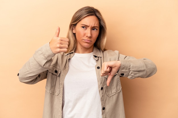 Giovane donna caucasica isolata su sfondo beige che mostra pollice in alto e pollice in basso, difficile scegliere concept