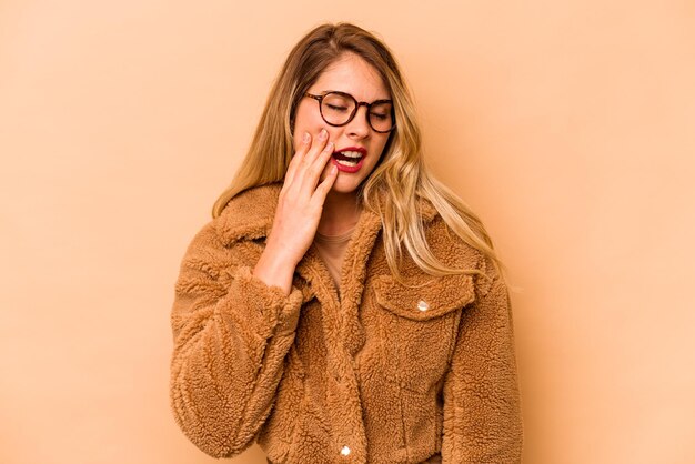 Giovane donna caucasica isolata su sfondo beige che sbadiglia mostrando un gesto stanco che copre la bocca con la mano