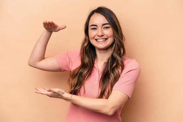 Giovane donna caucasica isolata su sfondo beige che tiene qualcosa di piccolo con gli indici sorridenti e fiduciosi