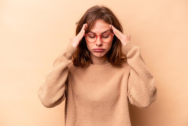 Giovane donna caucasica isolata su sfondo beige che tocca le tempie e ha mal di testa