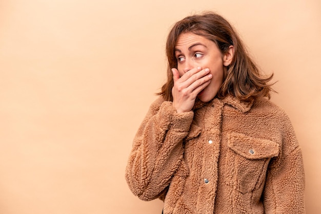 Giovane donna caucasica isolata su sfondo beige premurosa guardando uno spazio di copia che copre la bocca con la mano