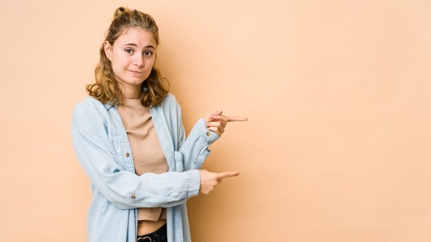 Giovane donna caucasica isolata su sfondo beige scioccato che punta con il dito indice a uno spazio di copia.