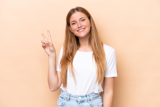 Giovane donna caucasica isolata su sfondo beige sorridendo e mostrando il segno di vittoria