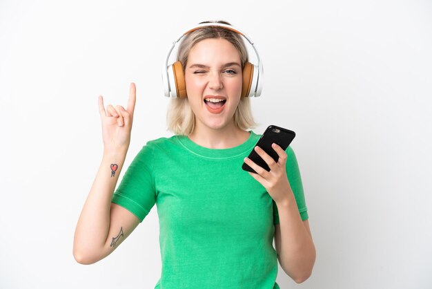 Giovane donna caucasica isolata su sfondo bianco ascoltando musica con un cellulare che fa gesto rock