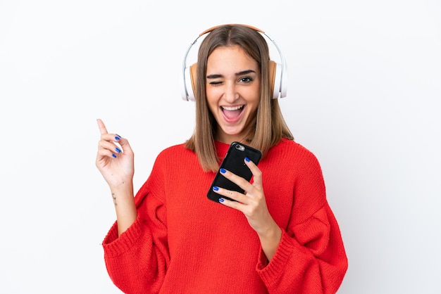 Giovane donna caucasica isolata su sfondo bianco ascoltando musica con un cellulare e cantando