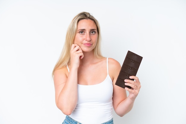 Giovane donna caucasica isolata su sfondo bianco prendendo una tavoletta di cioccolato e avendo dubbi