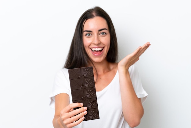 Giovane donna caucasica isolata su sfondo bianco prendendo una tavoletta di cioccolato e sorpresa