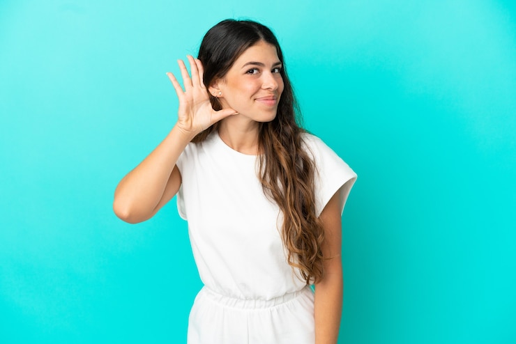 Foto giovane donna caucasica isolata su sfondo blu ascoltando qualcosa mettendo la mano sull'orecchio