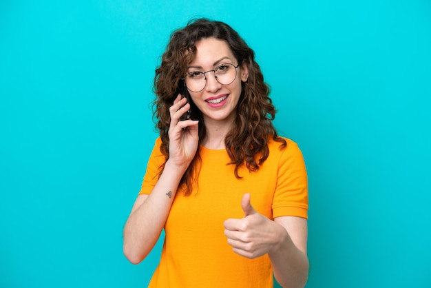 Giovane donna caucasica isolata su sfondo blu che tiene una conversazione con il cellulare mentre fa i pollici in su