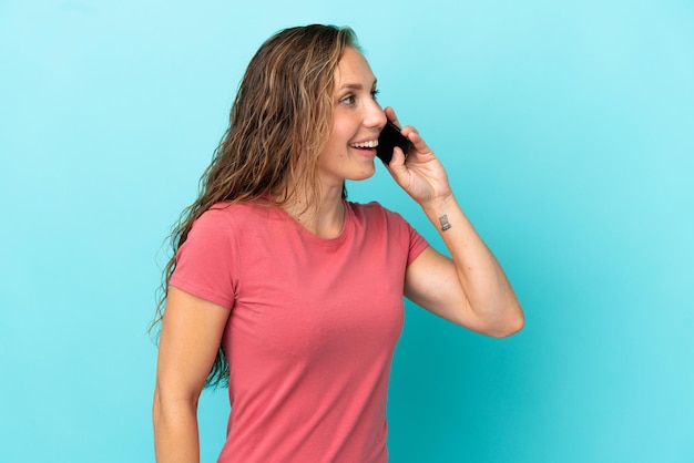 Giovane donna caucasica isolata su sfondo blu che tiene una conversazione con il telefono cellulare