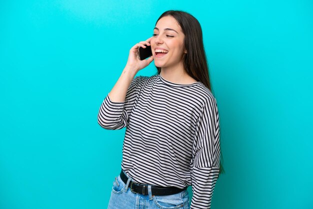 Giovane donna caucasica isolata su sfondo blu che tiene una conversazione con il telefono cellulare