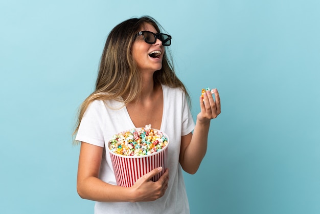 Giovane donna caucasica isolata su sfondo blu con occhiali 3d e con in mano un grande secchio di popcorn