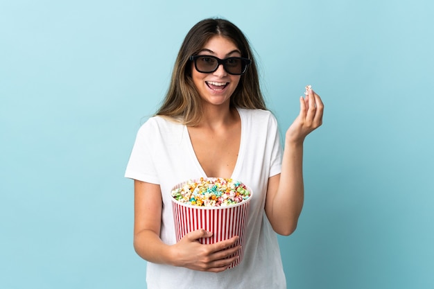 Giovane donna caucasica isolata su sfondo blu con occhiali 3d e con in mano un grande secchio di popcorn