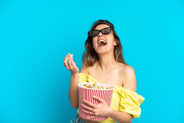 Giovane donna caucasica isolata su sfondo blu con occhiali 3d e con in mano un grande secchio di popcorn