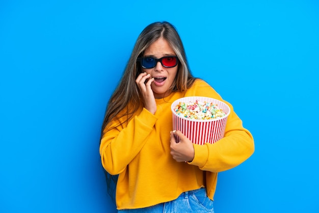 Giovane donna caucasica isolata su sfondo blu con occhiali 3d e con in mano un grande secchio di popcorn