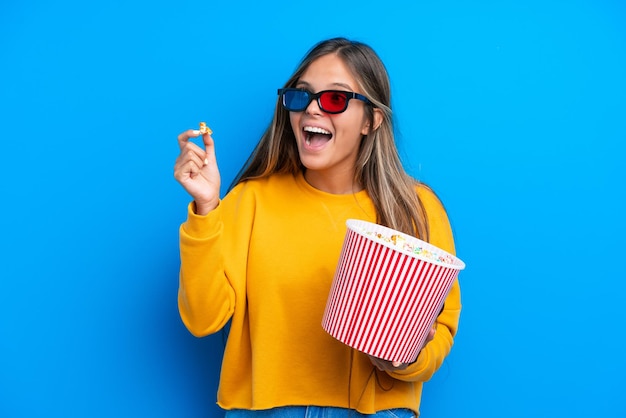 Giovane donna caucasica isolata su sfondo blu con occhiali 3d e in possesso di un grande secchio di popcorn