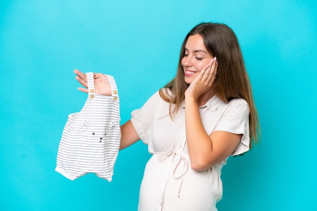 Giovane donna caucasica isolata su sfondo blu incinta e tenendo i vestiti del bambino