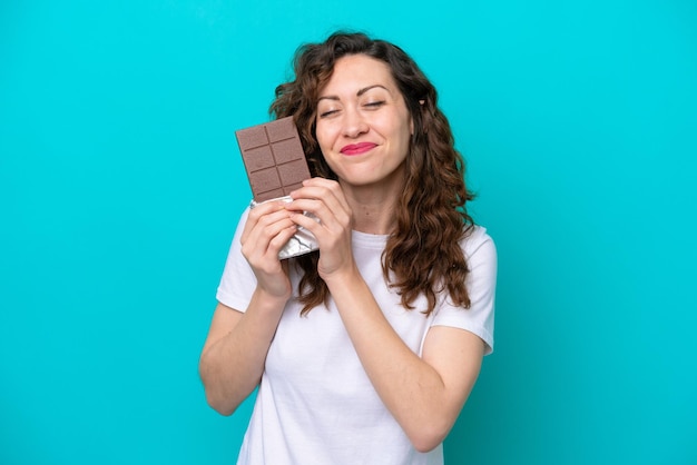 Giovane donna caucasica isolata su sfondo blu, prendendo una tavoletta di cioccolato e felice