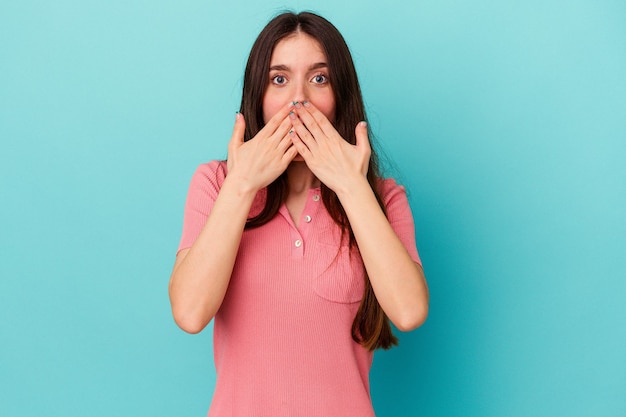 Giovane donna caucasica isolata su sfondo blu scioccata, coprendosi la bocca con le mani, ansiosa di scoprire qualcosa di nuovo.