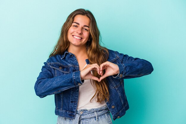 Giovane donna caucasica isolata su sfondo blu sorridente e mostrando una forma di cuore con le mani.