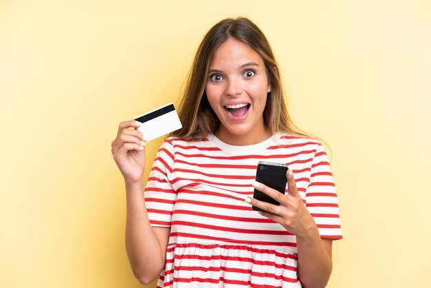 Giovane donna caucasica isolata su sfondo giallo, acquisto con il cellulare e in possesso di una carta di credito con espressione sorpresa
