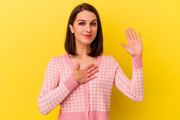 Giovane donna caucasica isolata su sfondo giallo che presta giuramento mettendo la mano sul petto