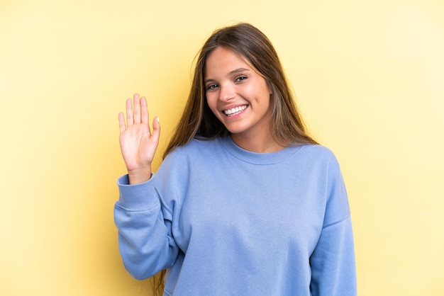 Giovane donna caucasica isolata su sfondo giallo che saluta con la mano con un'espressione felice