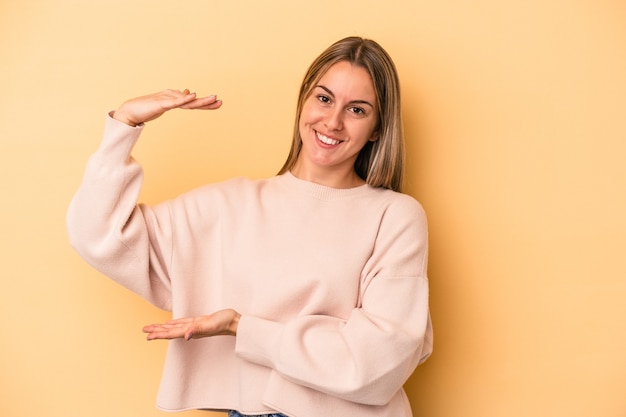 Giovane donna caucasica isolata su sfondo giallo che tiene qualcosa di piccolo con l'indice, sorridente e sicura di sé.