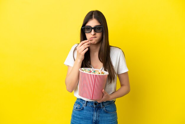 Giovane donna caucasica isolata su sfondo giallo con occhiali 3d e con in mano un grande secchio di popcorn