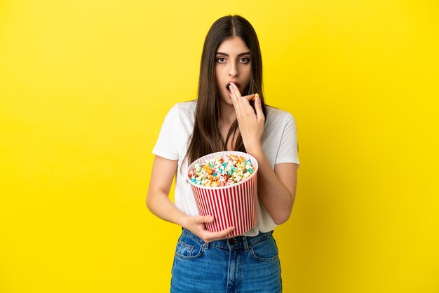 Giovane donna caucasica isolata su sfondo giallo in possesso di un grande secchio di popcorn