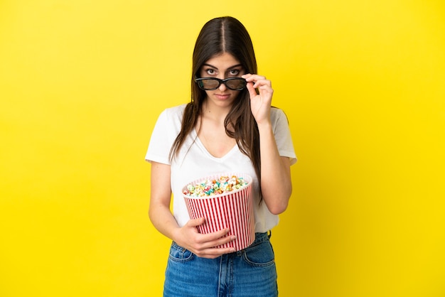 Giovane donna caucasica isolata su sfondo giallo sorpresa con occhiali 3d e con in mano un grande secchio di popcorn