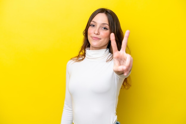 Giovane donna caucasica isolata su sfondo giallo sorridendo e mostrando il segno di vittoria