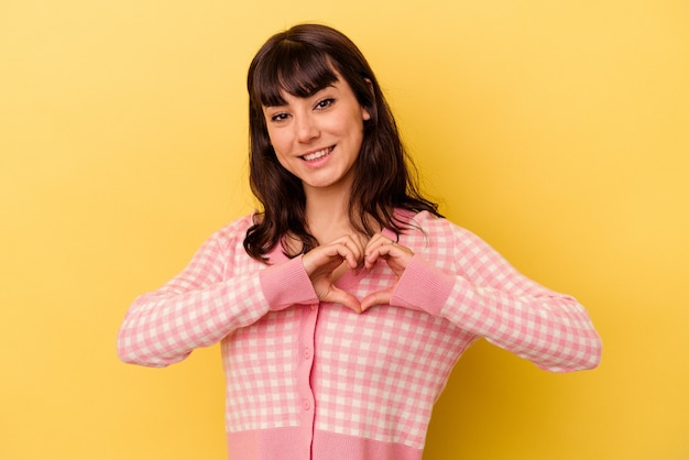 Giovane donna caucasica isolata su sfondo giallo sorridendo e mostrando una forma di cuore con le mani.