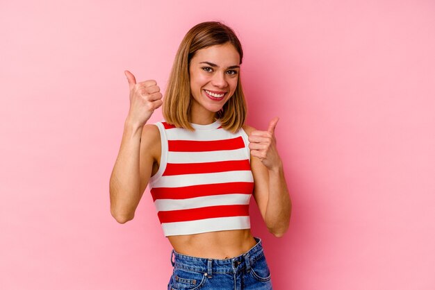 Giovane donna caucasica isolata su sfondo rosa, alzando entrambi i pollici in su, sorridente e fiduciosa.