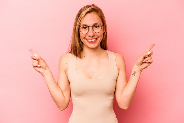 Giovane donna caucasica isolata su sfondo rosa che punta a diversi spazi di copia scegliendone uno che mostra con il dito