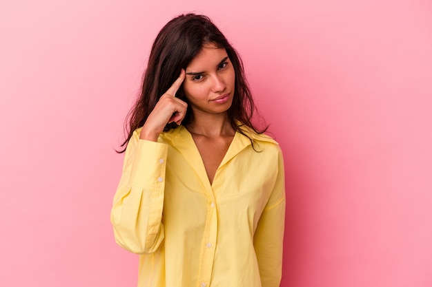 Giovane donna caucasica isolata su sfondo rosa che punta il tempio con il dito, pensando, concentrata su un compito.