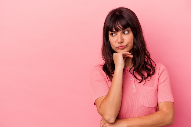 Giovane donna caucasica isolata su sfondo rosa guardando lateralmente con espressione dubbiosa e scettica.