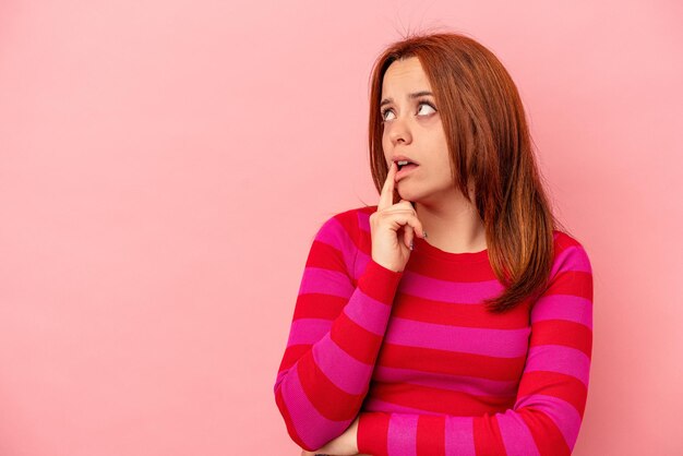 Giovane donna caucasica isolata su sfondo rosa guardando lateralmente con espressione dubbiosa e scettica.