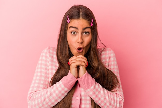 Giovane donna caucasica isolata su sfondo rosa pregando per la fortuna, stupita e aprendo la bocca guardando in avanti.